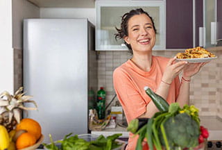 young woman it kitchen holding plate of food while smiling for camera